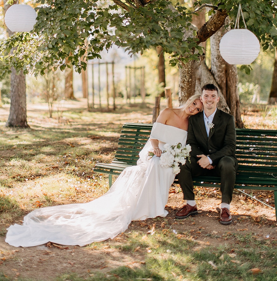 bride and groom sitting on a bench for a destination wedding in france bridal hair and makeup natural and brides hair in a bun with white lanterns hanging from the green summer trees