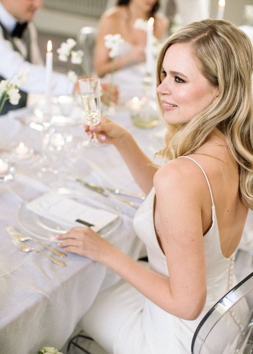 bride at the dinning table wearing sleek wedding dress, natural makeup and blonde hair down in a natural wave